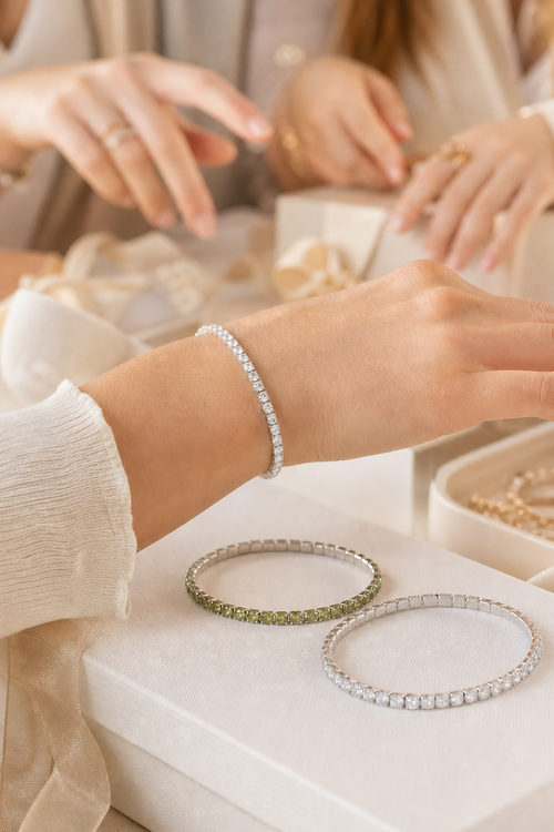 A close-up lifestyle shot of a person wearing a delicate silver tennis bracelet, with two other bracelets displayed on a white jewelry box. One bracelet is silver with green stones, and another is a classic silver tennis bracelet.
